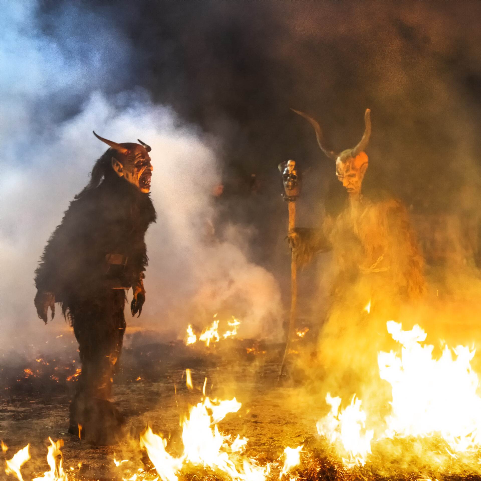 Zwei Perchten mit Hörnermasken und Fellkostümen zwischen Feuer und Rauch